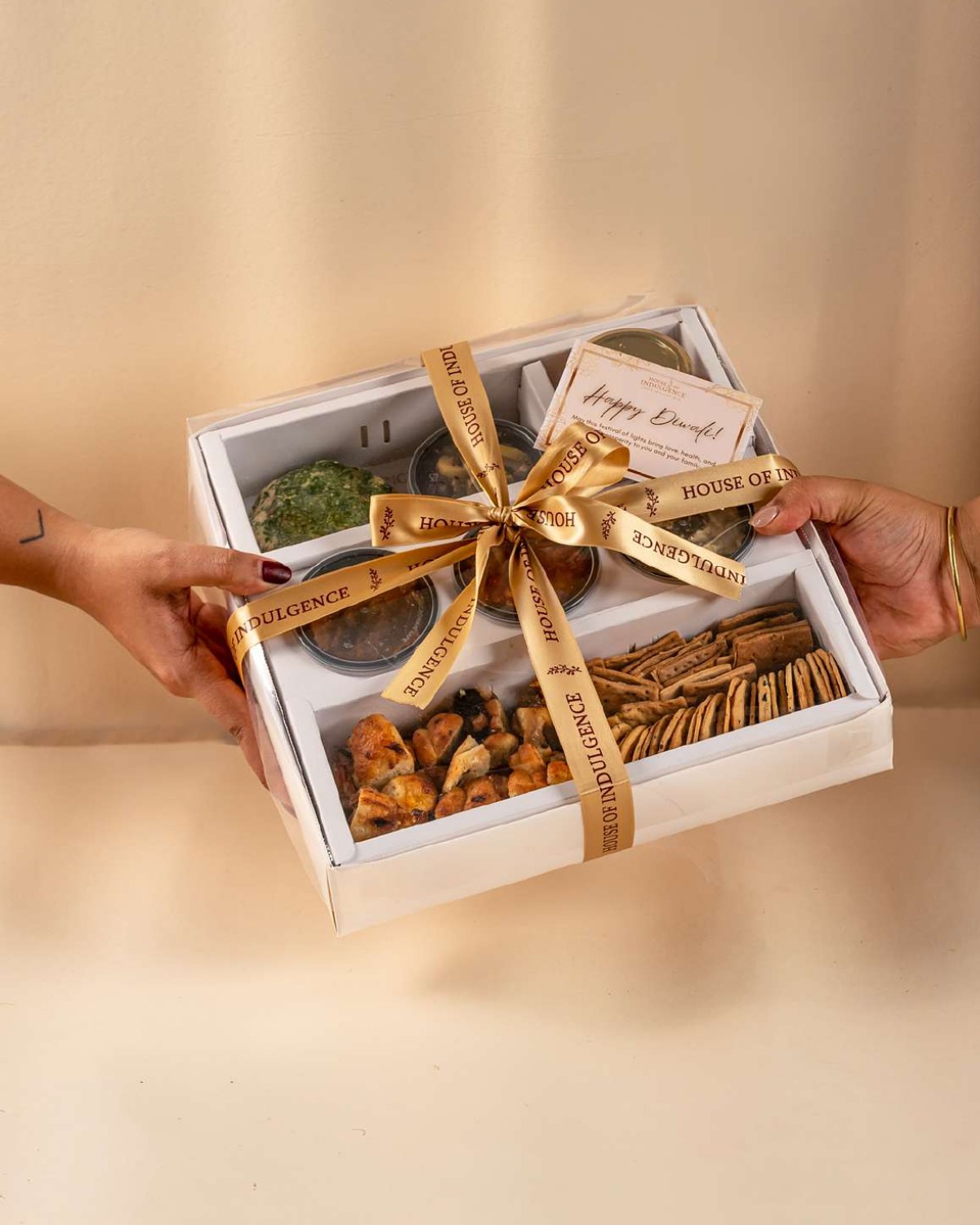 A House of Indulgence gift box with a herb-crusted cheese ball, dips, crackers and bread, tied with a gold ribbon and handed over